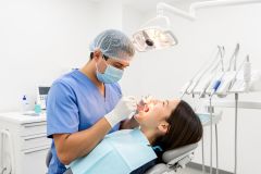 Dentist in scrubs and mask examines a patient's open mouth under a dental light in a sterile operatory.