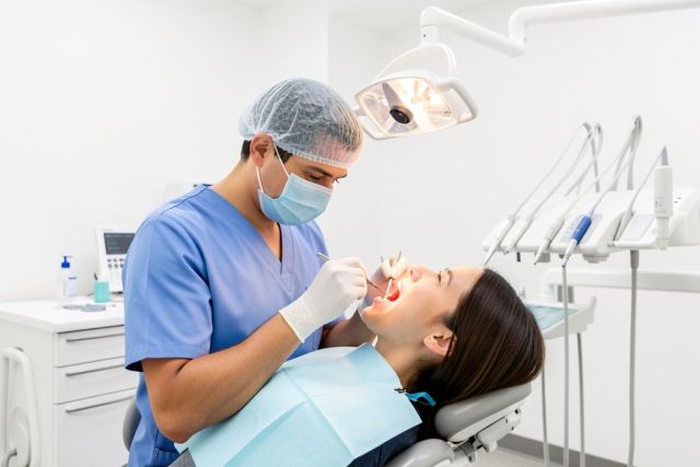 Dentist in scrubs and mask examines a patient's open mouth under a dental light in a sterile operatory.