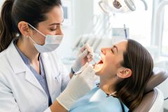 Dentist wearing mask and gloves examines a reclining patient's open mouth with mirror and probe in a bright dental clinic.
