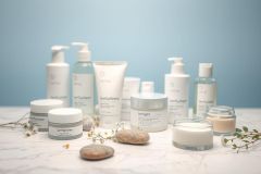 Product shot of Lumira skincare collection. White and silver containers featuring creams, lotions, and cleansers are arranged on a marble surface. Soft blue background creates a clean, spa-like aesthetic. Focus on natural beauty and luxurious skincare products.