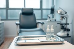Medical office interior featuring a black examination chair, stainless steel tray with dropper bottles, and medical equipment. The room has a clean, modern aesthetic with soft blue and white tones, and natural light from windows. Focus is on healthcare, dermatology, or cosmetic procedures within a clinical setting.
