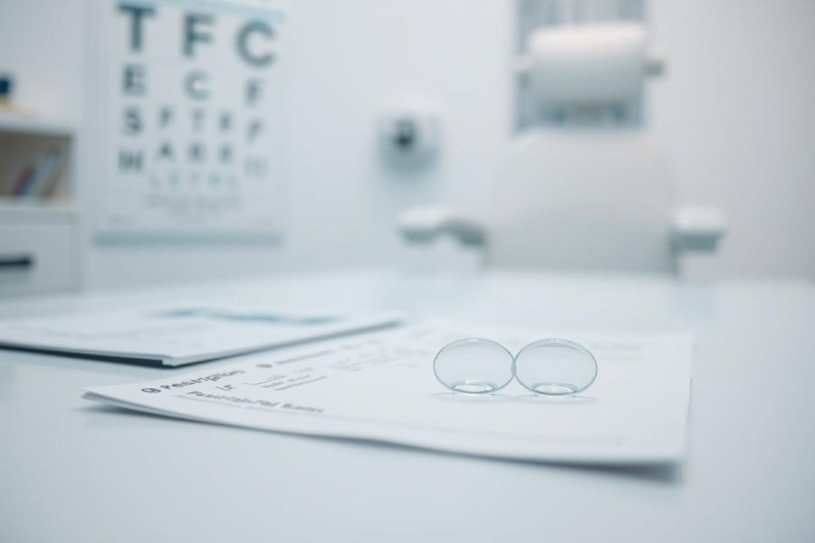 Close-up of clear glasses resting on eye exam paperwork in a bright, modern optometrist's office. Features an eye chart in the background, showcasing a healthcare and vision care setting. Predominantly white and blue tones.