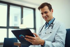 Doctor in white coat reviewing tablet, likely in a clinic or hospital setting. Focus on healthcare, medical technology, and professional environment. Blue shirt visible.