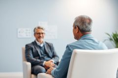 Two men in a professional setting, likely a therapy or consultation session. One man, seated and viewed from behind, wears blue. The other, in a suit, smiles warmly. The background is a soft blue with a blurred whiteboard, suggesting a calm and supportive environment. Keywords: therapy, consultation, men, business, healthcare, counseling.