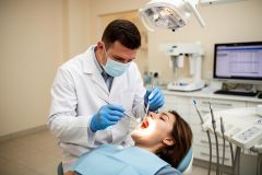 Dentist in mask examines patient's open mouth under a dental light during a clinical tooth exam.