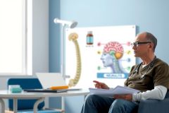 The image features a medical professional, likely a doctor or nurse, sitting in an office environment. The person is wearing glasses and has a laptop on the desk in front of them.