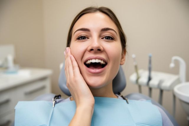 Young patient in a dental chair holding her cheek with mouth open, conveying dental discomfort or post‑treatment checkup.
