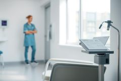 Hospital hallway scene featuring a nurse in blue scrubs standing in the background, an examination light over patient charts, and a glimpse of a medical bed. Clean, bright, and modern healthcare environment. Focus on medical care and patient documentation.