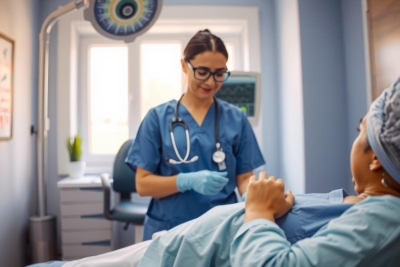 The image portrays a professional medical setting with a female doctor attending to a patient. The doctor is wearing blue scrubs and has a stethoscope around her neck, indicating her readiness for examination or diagnosis. She is standing next to an examination table where the patient is lying down, providing a sense of care and attention being given by the medical professional.
