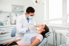 A dentist wearing mask and gloves examines a young patient's open mouth during a routine checkup.