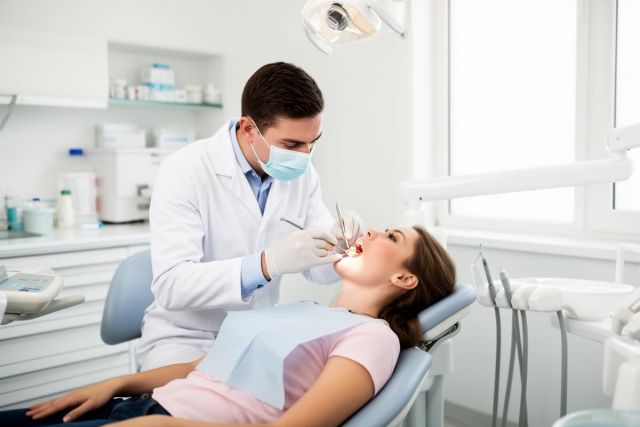 A dentist wearing mask and gloves examines a young patient's open mouth during a routine checkup.