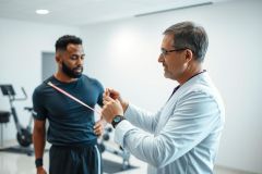 Doctor examining patient's shoulder in a gym setting. Male patient and physician, wearing a white coat, are visible. The image conveys a healthcare and fitness context with equipment in the background.