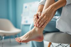 Woman experiencing ankle pain, sitting in a doctor's office. Close-up shot showcasing her foot and leg. Blue and neutral tones dominate the scene, suggesting a medical setting. Focus on foot discomfort and potential injury.