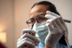 Dentist holding a dental tool, wearing a mask and gloves. Close-up of hands in a clinical setting, showcasing dental procedures and healthcare professionalism.