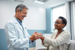 A caring doctor in a white coat comforts a patient in a bright, modern medical office. The image features a supportive interaction between a male physician and a female patient, emphasizing healthcare, empathy, and patient care in a clinical setting. Blue and white tones dominate the scene.
