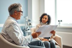 Doctor and patient consultation in a bright, modern clinic. A senior doctor examines medical documents while a young woman looks on, creating a healthcare discussion scene. Blue walls and natural light contribute to a professional and welcoming atmosphere.