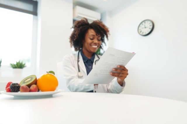 A healthcare professional in a white coat reviews a document at a table with fresh fruit. The scene suggests a focus on nutrition or patient care in a clinical setting.