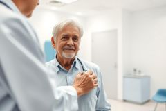 Senior man during a medical checkup in a bright, modern clinic. A doctor uses a stethoscope to listen to his heart. Healthcare, wellness, and preventative care themes are present. Light blue and white color palette.