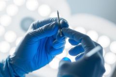 Close-up of gloved hands holding surgical instruments under bright lights. Focus on precision and medical procedure in a sterile environment. Blue background with bright illumination.