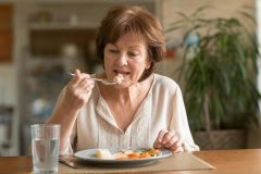 Elderly woman at dining table carefully chewing a small bite, plate, cutlery, and water glass visible.