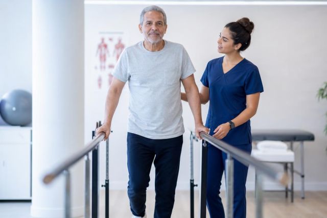 Older man performing parallel-bar walking exercise guided by a nurse in a rehabilitation clinic.