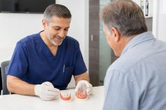 Dentist explains full denture and implant-supported prosthesis to older male patient in clinic.
