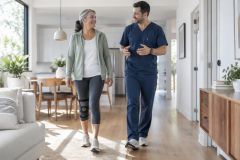 Older woman wearing knee brace walking at home with a smiling healthcare professional