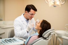 Dentist in protective eyewear examines a patient's teeth in a chair during a cosmetic dental consultation.