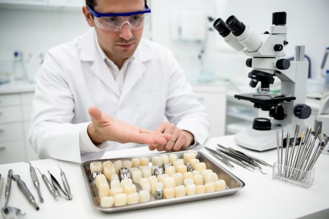 Dentist in a lab coat examining a tray of dental crown samples beside dental tools and a microscope