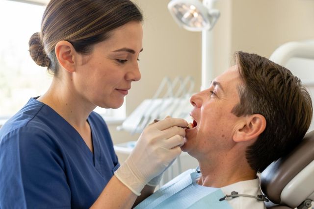 Dental clinician in scrubs holds a removable denture against a reclining patient's mouth during fit evaluation.