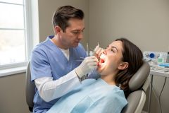A male dentist in blue scrubs conducts a professional dental examination on a female patient in a bright, modern clinic. The patient sits in a dental chair wearing a light blue bib while the dentist uses specialized tools for oral care. The clean, well-lit environment features medical equipment and a window, emphasizing a professional healthcare setting.