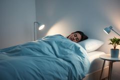 Woman sleeping peacefully in a modern bedroom. Soft blue bedding and a neutral color palette create a calm atmosphere. Two lamps illuminate the scene, showcasing a cozy and restful sleep environment.