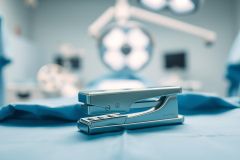 A silver stapler rests on a blue surgical drape in a blurred operating room setting. The image features medical equipment and blue lighting, creating a sterile and clinical atmosphere. Focus is on the stapler, suggesting office supplies in a healthcare context.