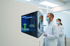 Scientists in lab coats examine a screen displaying 3D molecular structure. Sterile environment, likely a pharmaceutical research facility, with bright lighting and a focus on innovation.