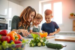 A mother and two children are preparing food together in a bright kitchen. Vegetables and fruits are on the counter. Focus on family cooking and healthy eating.