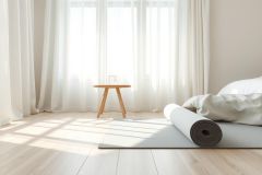 Bright, airy bedroom interior featuring a yoga mat and rolled yoga mat on a light wood floor. Soft, neutral colors—whites, creams, and beige—create a calming, minimalist atmosphere. A small wooden table sits beneath sheer white curtains and a window, suggesting a peaceful wellness space.