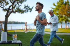 A man is jogging on a grassy area near water, with a bicycle and a leisurely setting that includes a drink with an orange slice. The environment suggests relaxation and outdoor activity. A physician in the background appears to be observing the runner.