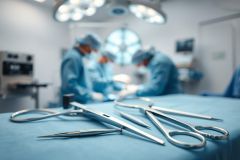 Surgical instruments, including forceps and scissors, rest on a blue surgical drape in a blurred operating room setting. Medical professionals in blue scrubs are visible performing surgery under bright overhead lights. Healthcare, surgery, and medical equipment themes are present.