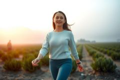The image depicts a woman jogging during sunset. The environment appears to be an outdoor, rural setting with a field of crops in the background.