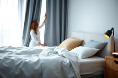 Woman adjusting curtains in a bright bedroom. Features a cozy bed with pillows, a bedside table with a lamp, and a minimalist aesthetic. Soft natural light and a calming color palette create a serene atmosphere.
