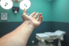 Close-up of a hand in a medical setting, likely an operating room. Surgical lights and equipment are visible in the blurred background. Focus on skin and healthcare context.