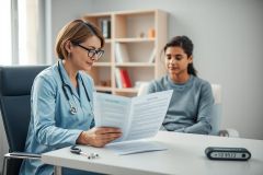 Doctor in a blue coat reviewing paperwork with a patient in a modern, bright medical office. Healthcare consultation, medical examination, and patient care scene. Focus on professional medical setting and doctor-patient interaction.