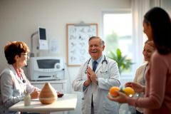 The image depicts a professional setting, likely a doctor's office or clinic. In the center stands a smiling male doctor wearing a medical gown and a stethoscope around his neck. He is surrounded by three female colleagues who are also dressed in medical attire. 