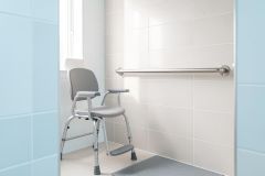 A grey medical shower chair with armrests and a footrest sits in a bright bathroom with white tiled walls and a silver safety grab bar.