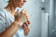 Woman scratching her arm, likely experiencing itchy skin. Soft, neutral tones dominate the image with a blurred bathroom background. Suggests skin irritation, allergies, or dry skin concerns.
