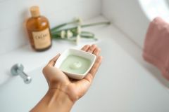 Close-up of a hand holding a bowl of green face mask, likely in a bathroom setting. Focus on skincare, self-care, and wellness. Features a ceramic bowl, white surfaces, and a natural, spa-like aesthetic.