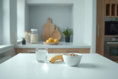Bright kitchen scene with a bowl of cereal, a banana, and a glass of water on a white countertop. Natural light highlights the breakfast setup, with wooden cabinets and plants in the background.