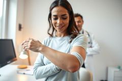 Woman examining her arm in a doctor's office. A healthcare professional stands in the background. The scene features a bright, clean interior with neutral colors and a focus on skin health and medical consultation.
