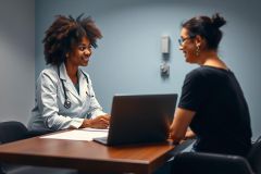 Doctor consults with patient in a modern office setting. Healthcare professional in white coat discusses concerns with a woman at a desk with a laptop. Blue walls and warm lighting create a professional atmosphere.