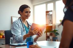 Doctor examining patient's medical record in a brightly lit office. Healthcare professional in white coat reviewing paperwork during consultation. Warm lighting, modern setting.